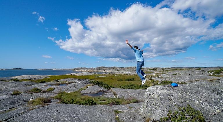 En person hoppar från en klippavsats mot en blå och molnig himmel.
