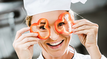 smiling female chef in double-breasted jacket holding pepper slices in front of face in restaurant kitchen