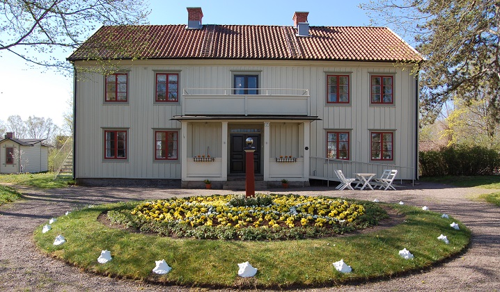 En stor vit träbyggnad i två våningar och med röda fönster. Framför byggnaden finns en rund rabatt, planterad med gula blommor och kantad med stora vita snäckor.