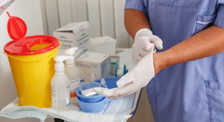 Närbild på en sköterska i blå arbetskläder som står vid ett provtagningsbord och tar på sig plasthandskar.