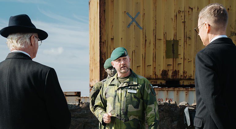 Den svenske kungen och den finske presidenten sedda bakifrån när de står och lyssnar till överste Fredrik
Herlitz i militäruniform. I bakgrunden en gul container. 