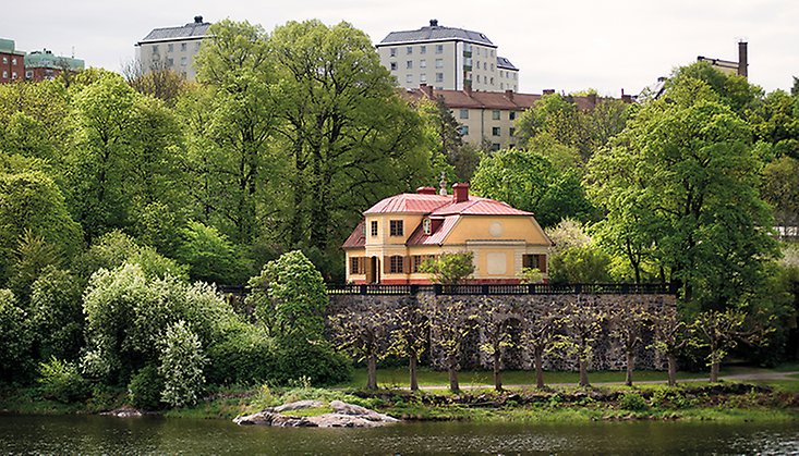 En 1700-tals byggnad vid vattnet omgiven av lummig grönska och med höghus i bakgrunden.