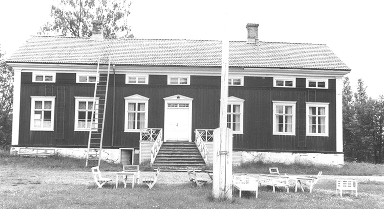 Gammal bild av trähus med tegeltak. Flaggstång samt bord och stolar står framför huset.
