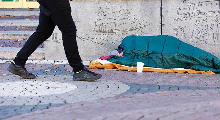 Person som ligger i en sovsäck på en trottoar. En vit pappmugg står framför.