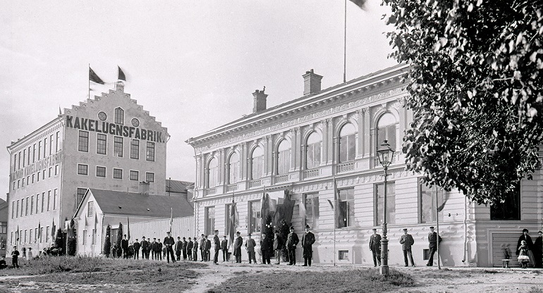 Gammal svartvit foto som visar kakelmakarens hus och kakelugnsfabriken i bakgrunden. Framför husen står många människor huvudsakligen mörkklädda män.