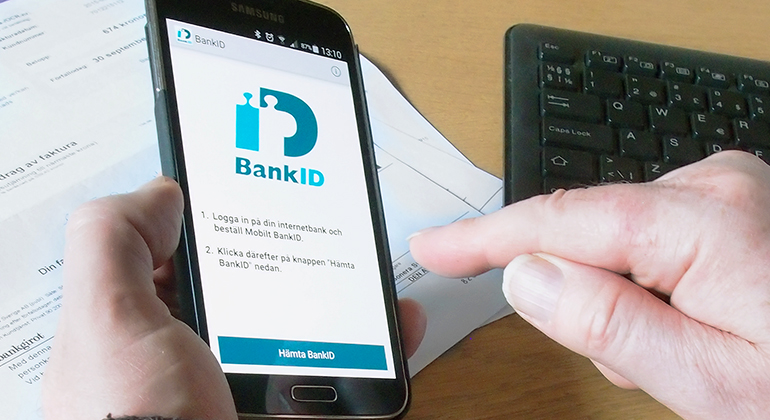 Närbild på händer som håller i en mobiltelefon där Bank-ID syns på skärmen. På bordet under syns ett tangentbord och en pappersfaktura.