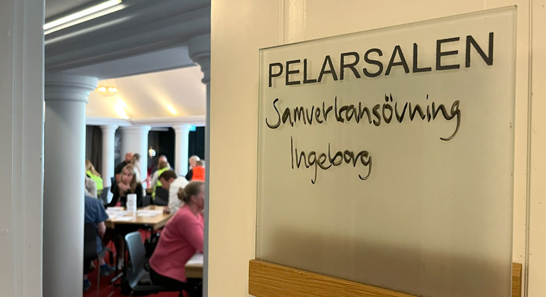 Närbild på en skylt utanför en lokal. På skylten står texten "Pelarsalen, samverkansövning Ingeborg". I bakgrunden skymtar ett rum där flera personer sitter runt olika bord.