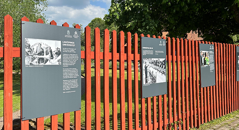 Skyltar med foton sitter upphängda på ett rött staket utanför lummig slottsträdgård.