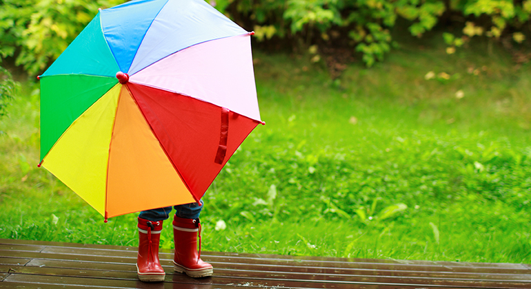 Ett barn i gummistövlar står gömd bakom ett färgglatt paraply i regnet.