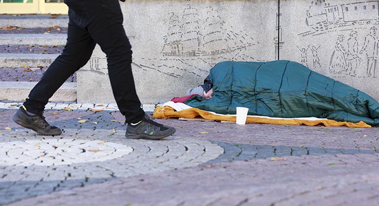 Person som ligger i en sovsäck på en trottoar. En vit pappmugg står framför.