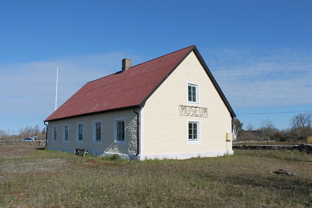 En ljust gul byggnad i sten med ett rött plåttak ligger på en tomt som avgränsas av en låg stenmur. Byggnaden ligger på en öppen gräsbevuxen tomt om omgärdas av en låg kalkstensmur.