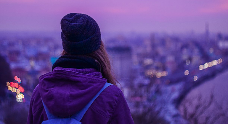 En person med långt hår, mössa, halsduk, vinterjacka och ryggsäck står med ryggen mot kameran och blickar ut över en stad i blått skymningsljus.