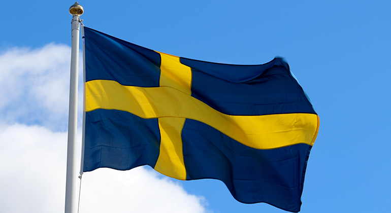 Svenska flaggan vajar på sin stång mot en blå himmel. 