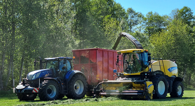Jordbruksmaskiner i arbete på gräsfält med lövskog i bakgrunden.