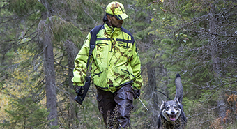 En jägare med reflekterande jaktkläder vandrar i skogen tillsammans med en grå jakthund. 