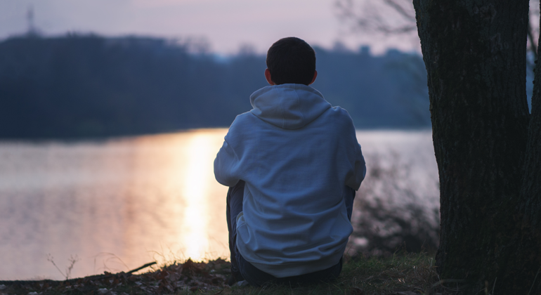En tonårspojke sitter ensam och tittar ut över en sjö. 