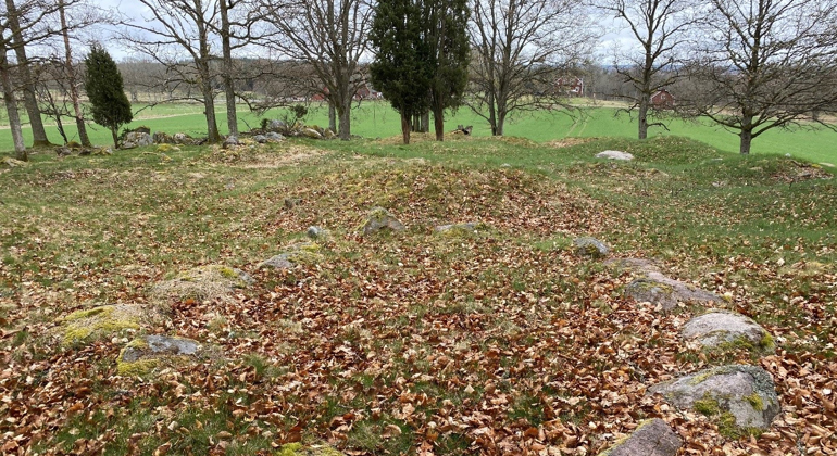 En ring med stenar jämnt utplacerade formar en grav. I bakgrunden skymtar gravhögar innan åkermark och gårdsbebyggelse tar vid.