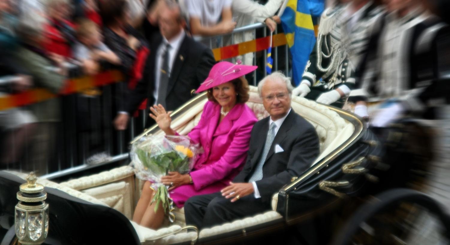 Bild på kungen och drottningen i öppen hästvagn, drottningen i rosa dräkt och kungen i kostym. Suddig omgivning. 