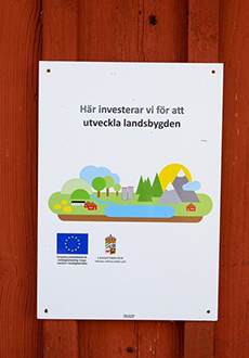 En skylt sitter uppsatt på en röd trävägg. Skylten har texten "Här investerar vi för att utveckla landsbygden" samt EU-logga och Länsstyrelsen Västra Götalands logga.