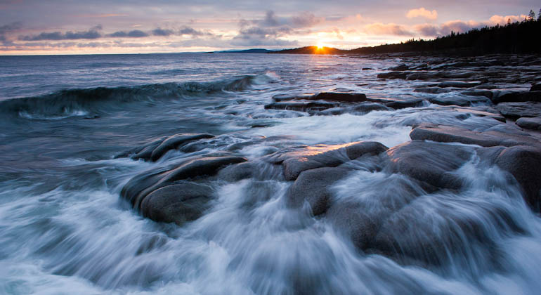 Foto av hur vågor rinner över klipporna i solnedgång.