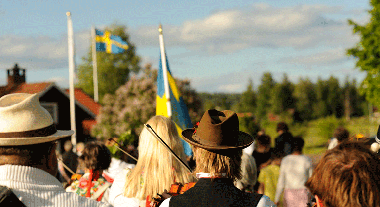 Ryggen på spelmän och kvinnor syns i förgrunden. Bakom syns svenska flaggan hissad. 