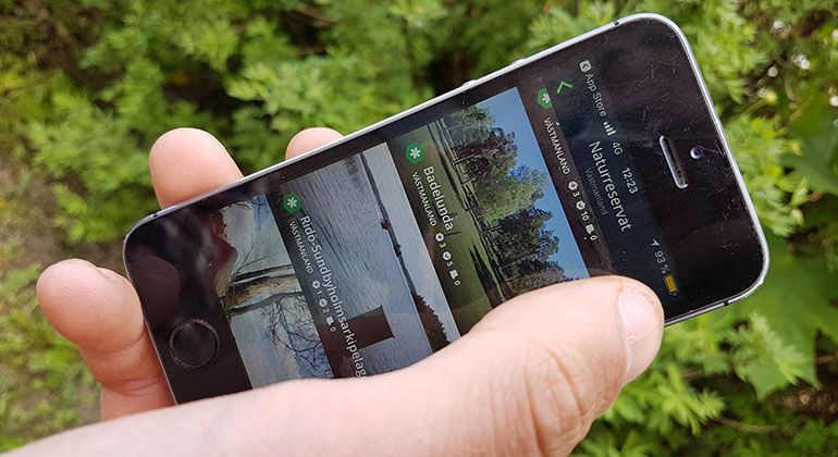 En telefon med appen Naturkartan öppen, en hand med en tumme som scrollar på telefonen.