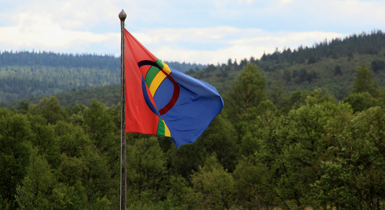 samiska flaggan som vajar i vinden. Bakom ses skogsvidder på kuperad mark mot himmel.
