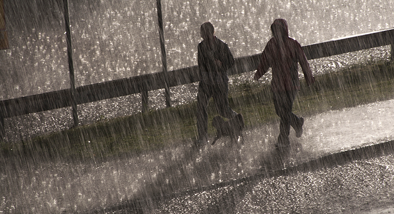 Vi ser två personer som vandrar på en trottoar genom ett skyfall. Personerna har regnkläder på sig och vi kan inte se deras ansikten för kläderna och regnet. 