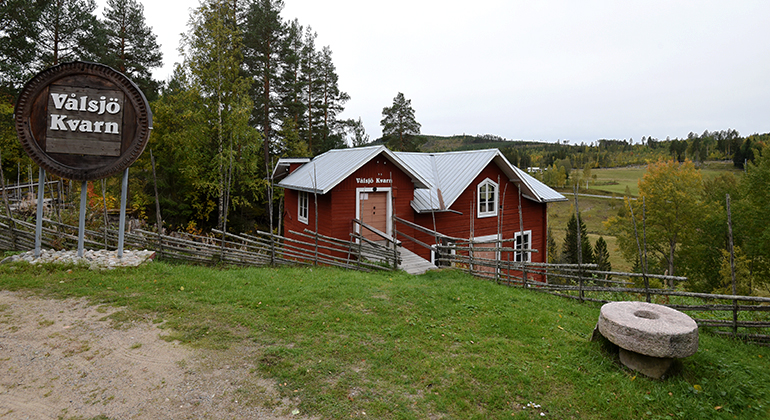 Entréskylt till Vålsjö kvarn. Bakom den finns en gärdsgård och en röd byggnad. Längst till höger i bilden - en kvarnsten.