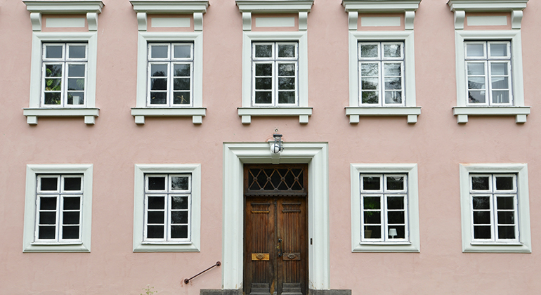 Närbild på huvudentrén där man ser två av husets våningar med fönster och den bruna ytterdörren.