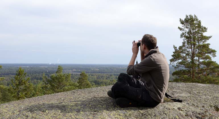 På en rundad häll med utsikt sitter en person och tittar med kikare ut över landskapet