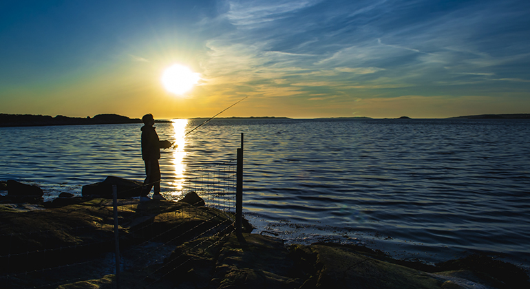 Foto taget i motljus på fiskare som står på en sten och fiskar i havet. 