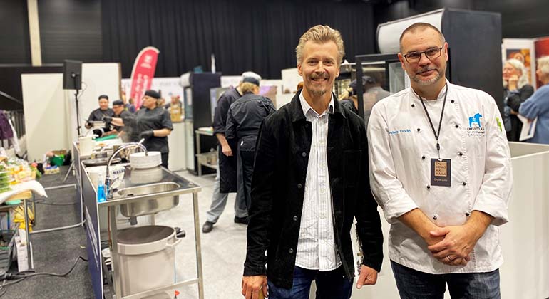 Paul Svensson och Anders Frodig tittar rakt in i kameran. I bakgrunden kockar som lagar mat. 