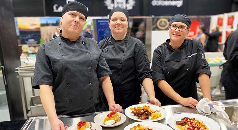 Tre kvinnor, vinnarna av kocktävlingen, kollar in i kameran. Framför dem står deras maträtter. 