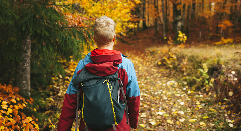 En person med kort ljust hår vandrar med ryggsäck i en höstig skog med färgglada löv på marken.