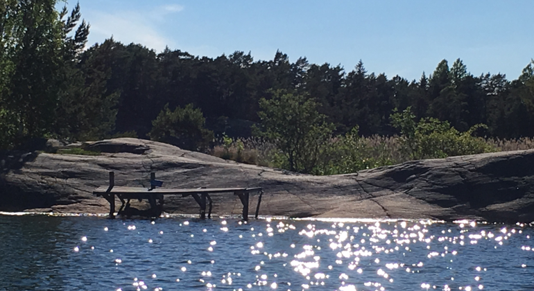 Skog, släta klipphällar, brygga och solglitter i vattenytan i Stavsudda-Krokholmens naturreservat. Foto: Länsstyrelsen