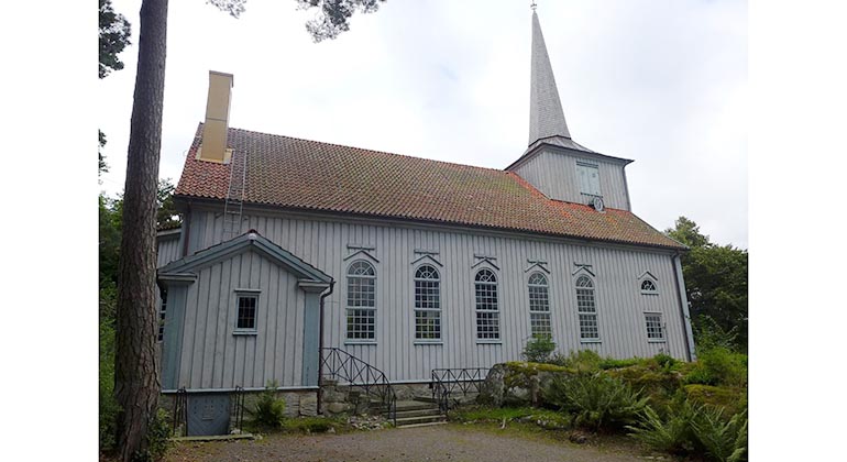 En grå träkyrka med ett spetsigt torn. Kyrkan har fem höga smala fönster.