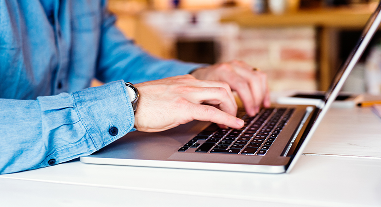 Fotot visar överkroppen av en person i jeansskjorta som sitter och arbetar vid en laptop i hemmiljö.