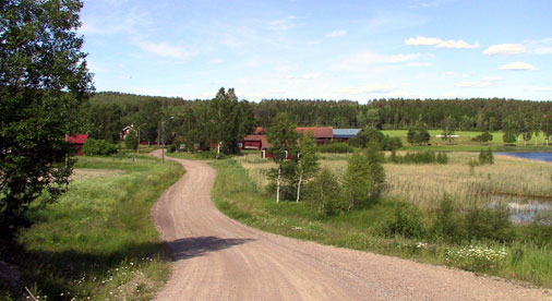 En väg som slingrar sig fram genom ett landskap, på sidan av vägen står dalaröda hus