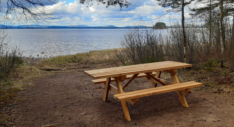 Rastbord med bänkar vid strandkant med en stor sjö i bakgrunden.