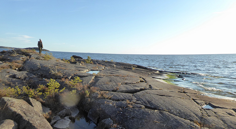Långt ut på de karga klipporna står en människa och tittar ut över det glittrande vattnet.