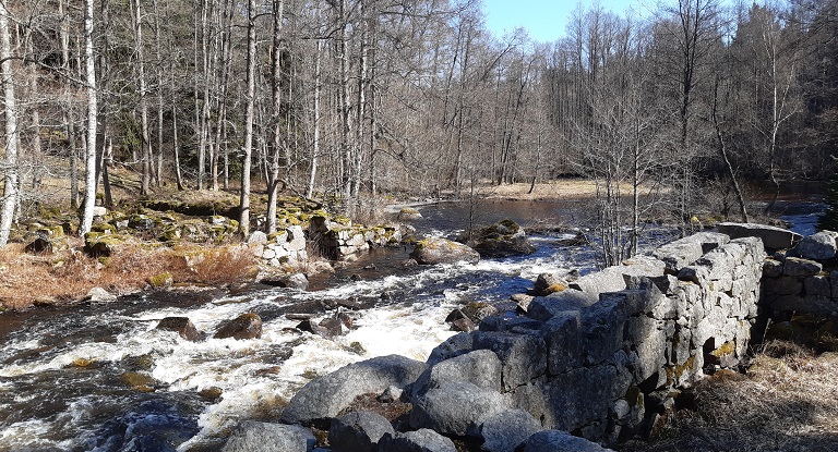 Bild på ett forsande vattendrag som rinner genom lövskog med kala träd. Höga stenmurar finns på båda sidor om vattnet, vilka utgör ruiner efter tidigare pappersbruk.