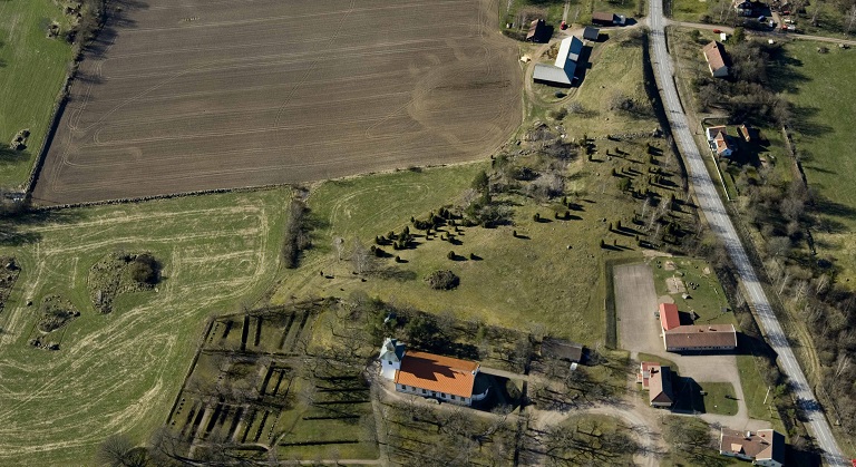 Ett foto från luften över ett öppet landskap där en kyrka och några gårdar syns. I högra kanten sträcker sig en landsväg.