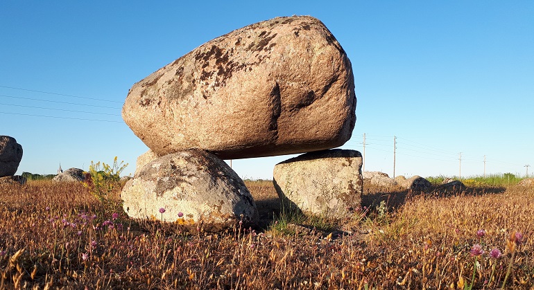Bild på en stor sten som placerats på tre mindre i en gräsmark. Fler liknande stenpyramider syns i bakgrunden.