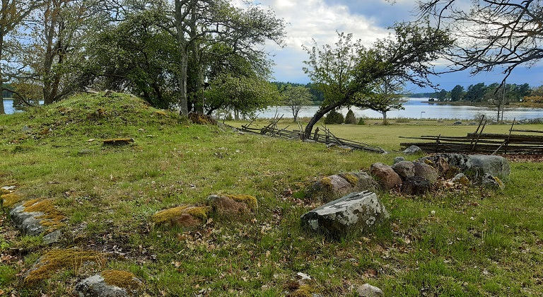 Foto på gräsklädd mark med träd och med öppet vatten i bakgrunden. I marken syns husgrund med stora stenar och en liten gräsklädd kulle, troligen röse efter spis.