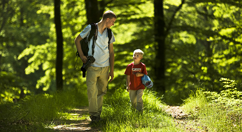 En pappa och son är ute och promenerar i naturen. I bakgrunden ser vi grönskkande träd och pappan har en kamera som hänger på ena axeln. 