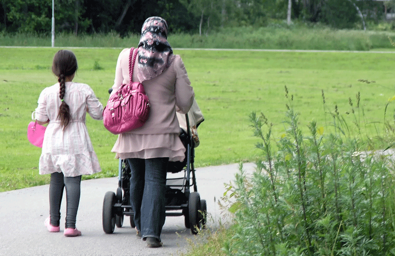 En mamma med en barnvagn och en flicka som går bredvid går på en promenadvagn. Familjen syns bakifrån. 