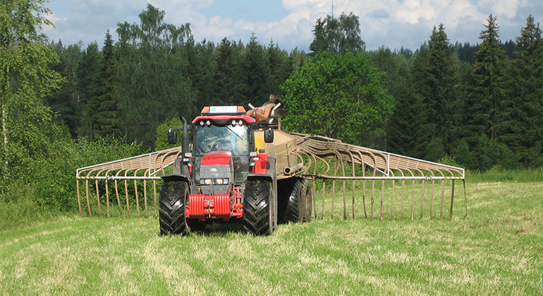 Röd traktor sprider stallgödsel på åkermark. Skog i bakgrunden.
