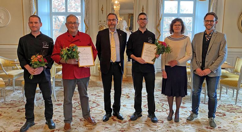 Sex personer  på rad i hörnsalongen i landshövdingens residens på Västerås slott. Tre av personerna håller i diplom och blommor för att de tagit fram Västmanlands landskapsrätter.