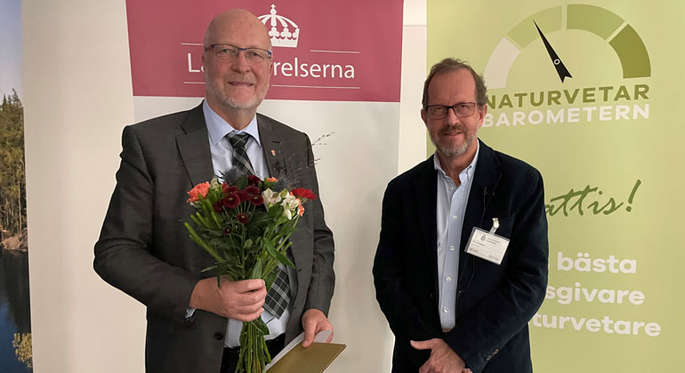 Sven-Erik Österberg, landshövding i Stockholms län, står med diplom och blommor i händerna. Bredvid står Erik Aronsson från fackförbundet Naturvetarna. 